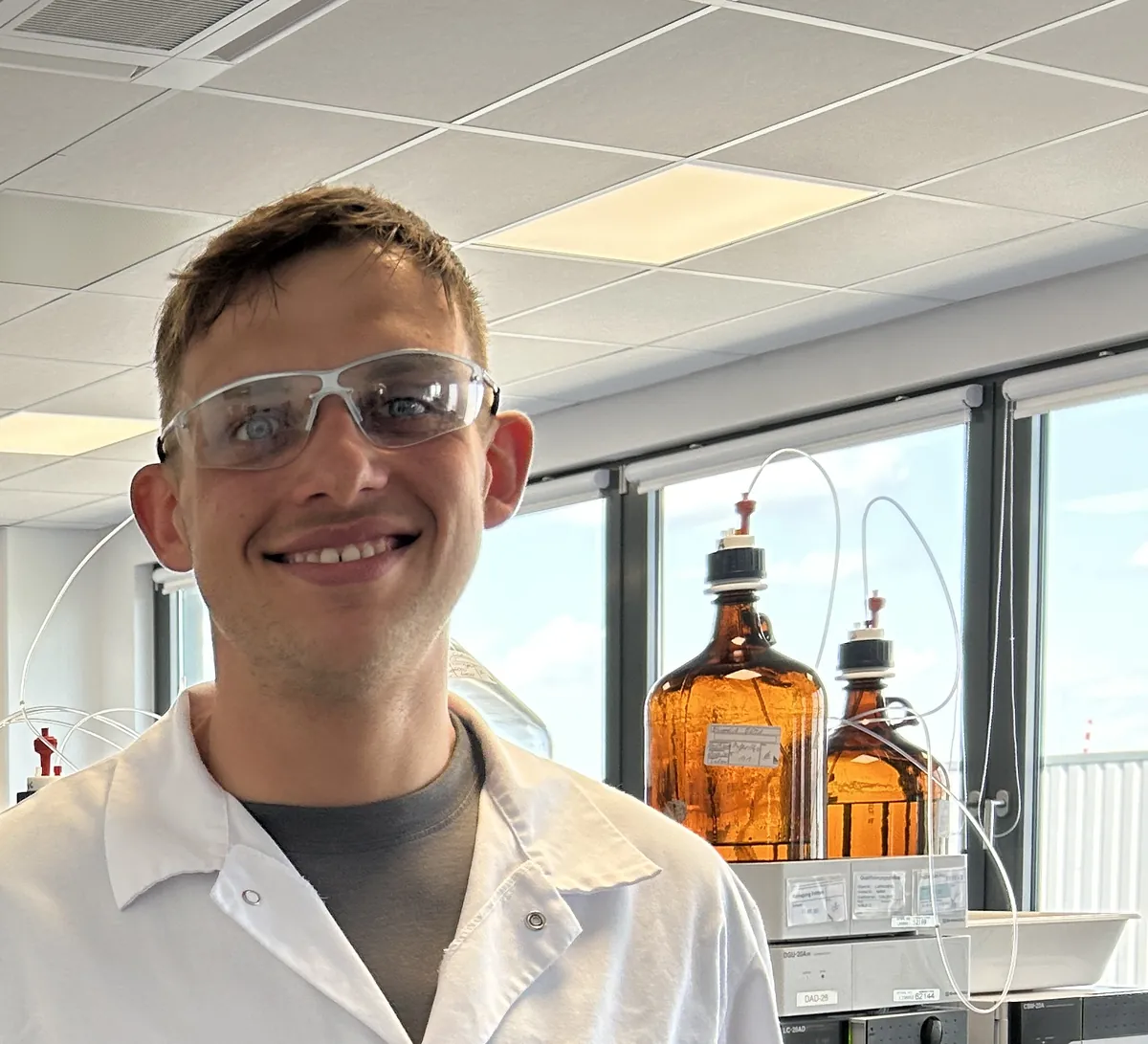 Peter Maibaum lacht mit Schutzbrille im Laborkittel, hinter ihm HPLC-Anlagen