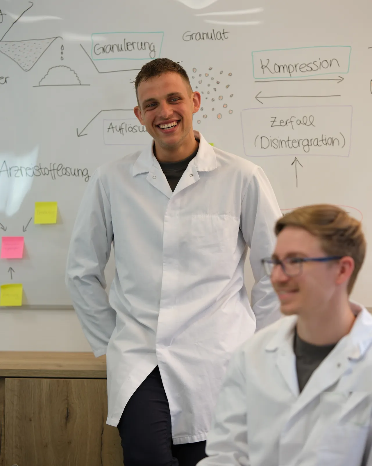 Peter Maibaum im Laborkittel vor einem Whiteboard mit Skizzen zur Tablettenherstellung, daneben ein Auszubildender